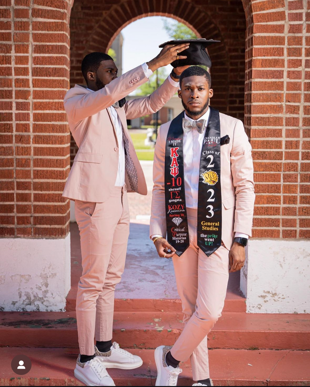 Graduate in formal attire with graduation caps and sashes in front of a brick building.