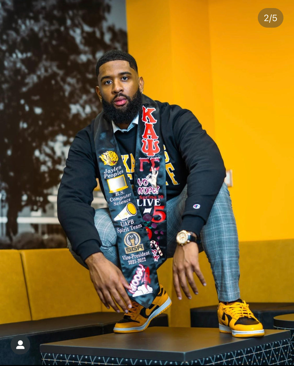 Man wearing a colorful scarf with text and logos, sitting on a platform against a yellow wall.