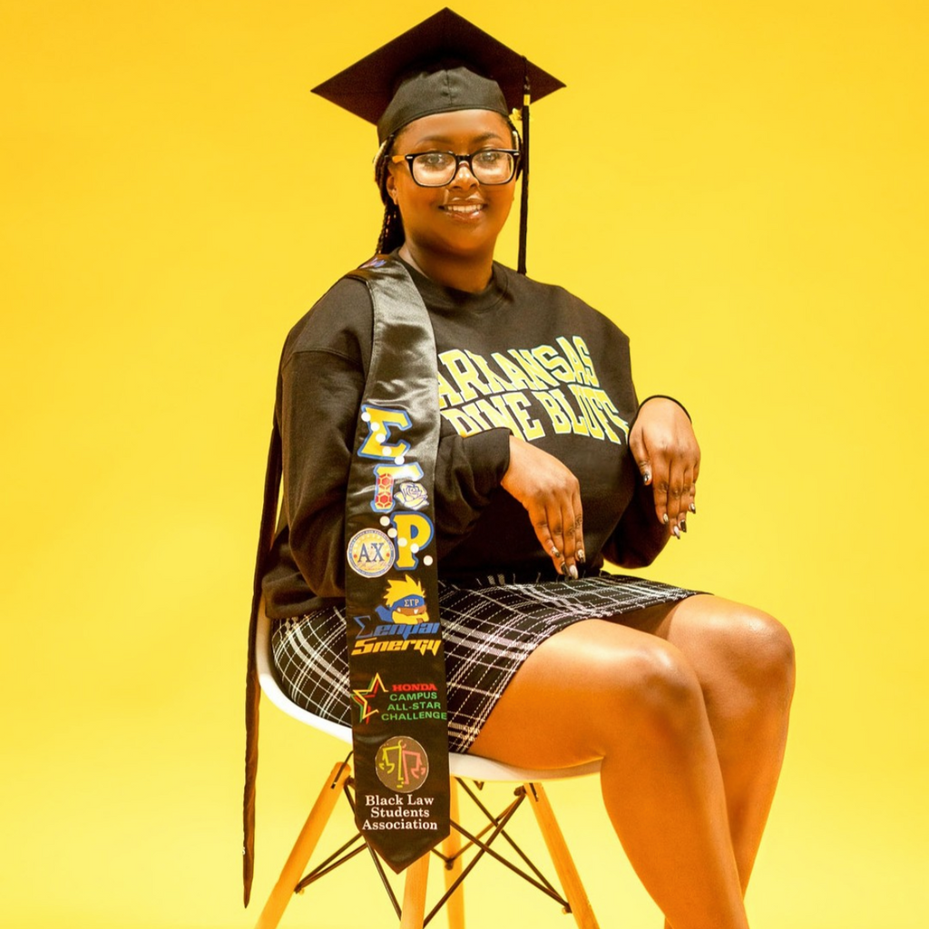 Graduate in cap and gown with a sash on a yellow background